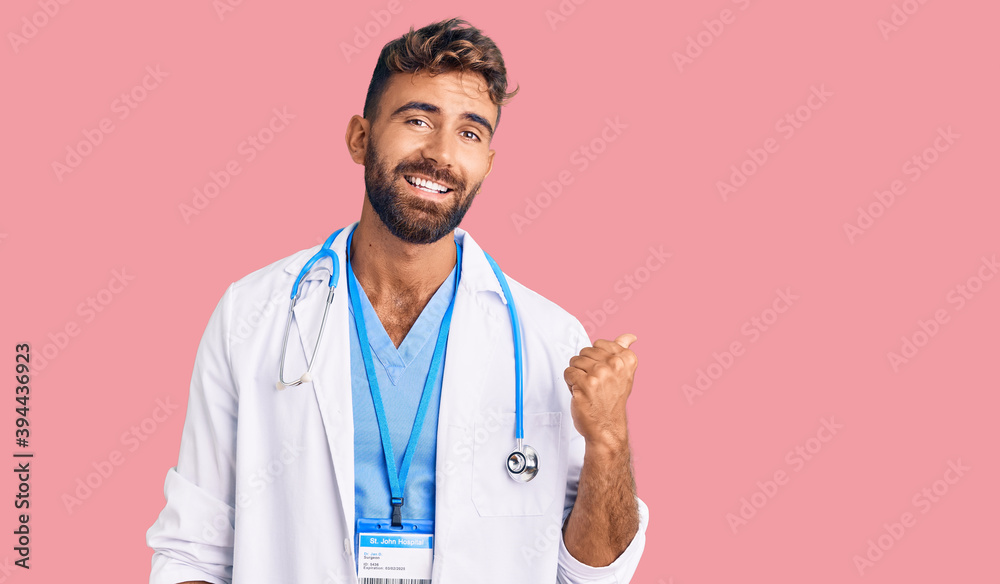Young hispanic man wearing doctor uniform and stethoscope smiling with happy face looking and pointing to the side with thumb up. Young hispanic man wearing doctor uniform and stethoscope smiling with happy face looking and pointing to the side with thumb up.