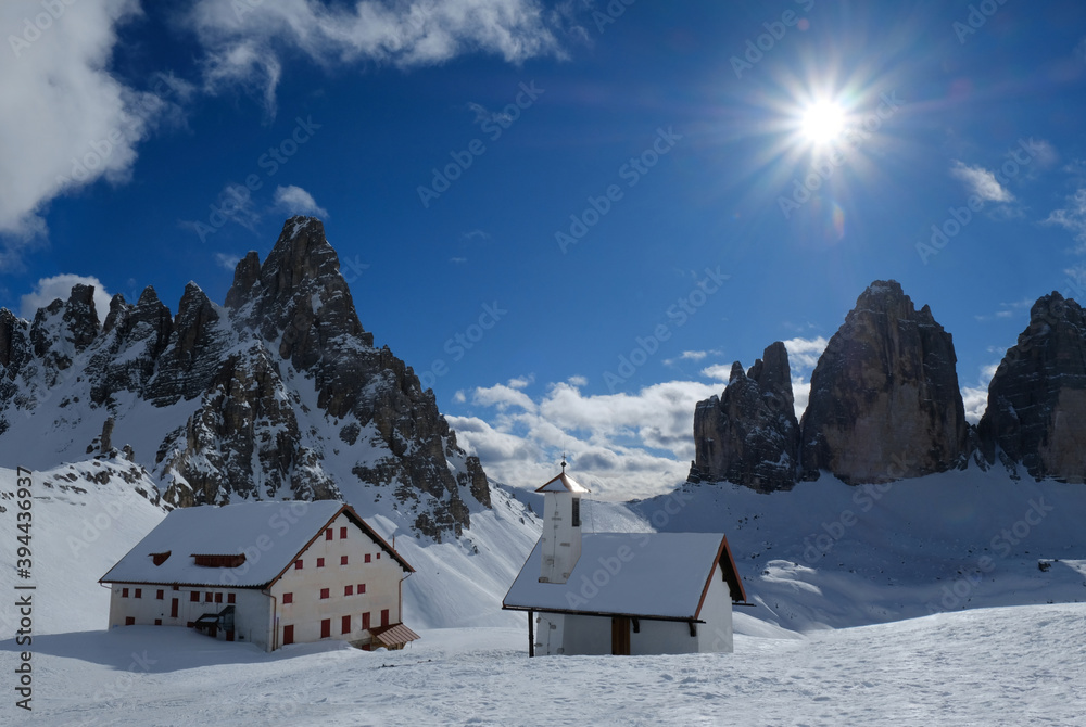 Beautiful panorama of Tre Cime di Lavaredo and Monte Paterno in sunny winter scenery. Refuge and chapel at foot of mountains. Dolomites, Italy