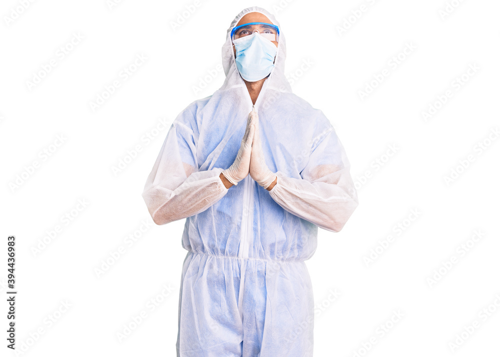 Young hispanic man wearing doctor protection coronavirus uniform and medical mask begging and praying with hands together with hope expression on face very emotional and worried. begging. Young hispanic man wearing doctor protection coronavirus uniform and medical mask begging and praying with hands together with hope expression on face very emotional and worried. begging.