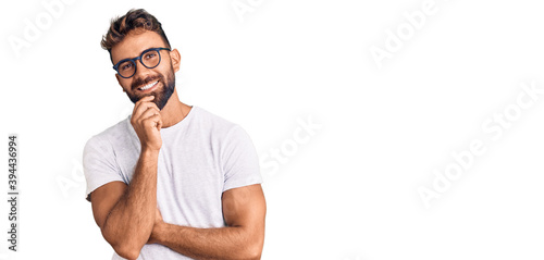 Young hispanic man wearing casual clothes and glasses looking confident at the camera smiling with crossed arms and hand raised on chin. thinking positive.