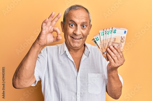 Handsome mature man holding bunch of 50 euro banknotes doing ok sign with fingers, smiling friendly gesturing excellent symbol photo