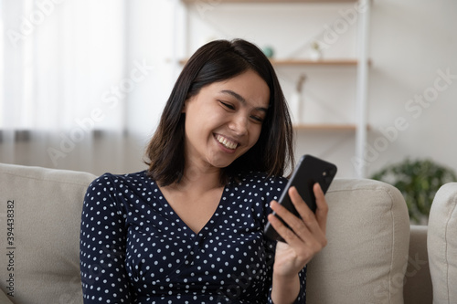 Close up smiling Asian young woman looking at phone screen, sitting on cozy couch at home, happy attractive female chatting or shopping online, having fun with mobile device, enjoying weekend