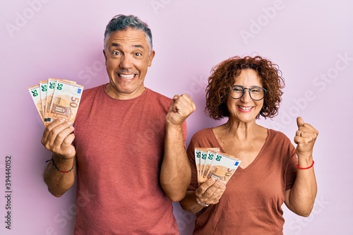 Beautiful middle age couple holding bunch of 50 euro banknotes screaming proud, celebrating victory and success very excited with raised arm