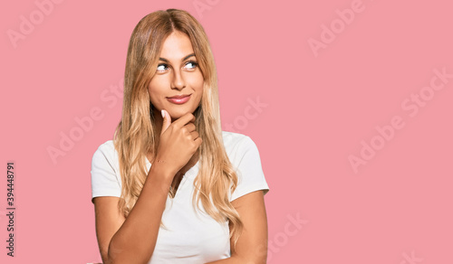 Beautiful blonde young woman wearing casual white tshirt with hand on chin thinking about question, pensive expression. smiling with thoughtful face. doubt concept.