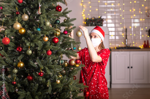 Young woman wearing face mask and pyjamas decorating christmas tree at home alone. celebrating xmas amnd new year during lockdown and pandemic photo