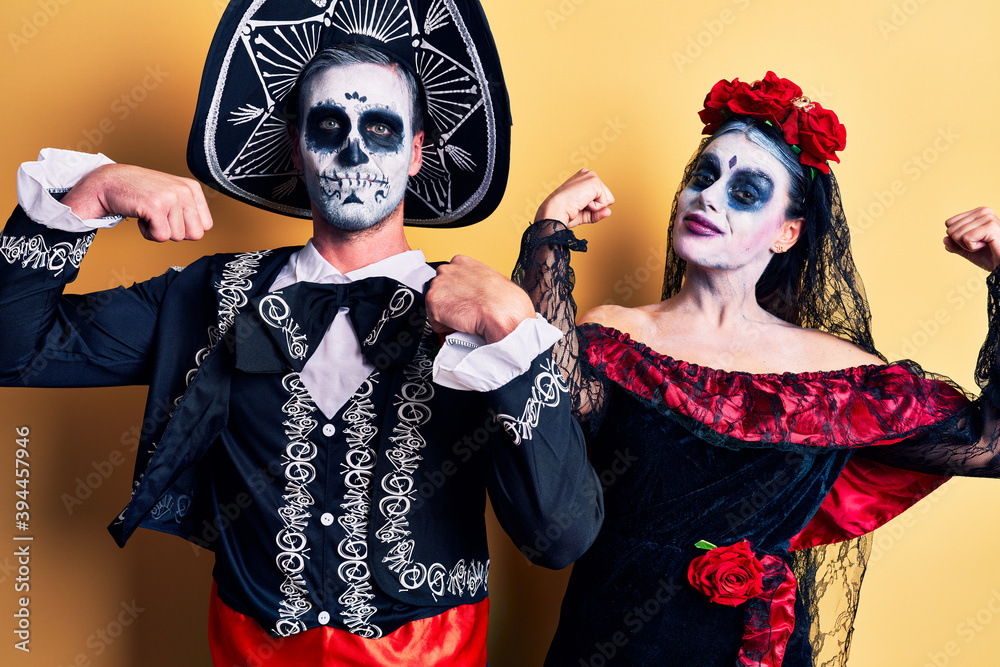 Young couple wearing mexican day of the dead costume over yellow showing arms muscles smiling proud. fitness concept.