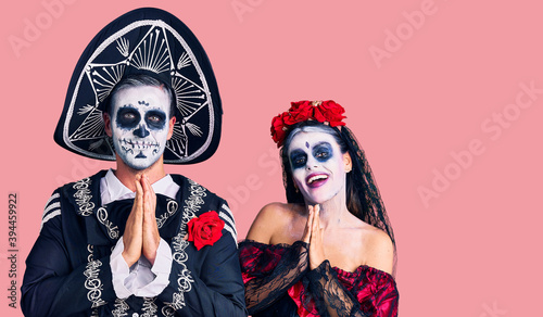 Young couple wearing mexican day of the dead costume over background praying with hands together asking for forgiveness smiling confident.