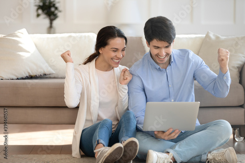 That is great. Overjoyed young husband and wife spending free time together resting on warm floor at home with laptop screaming in delight cheering for favorite sport team receiving good news by email