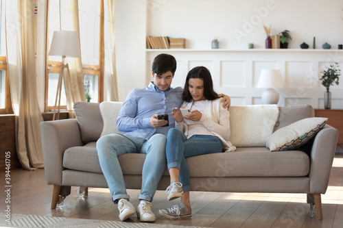 Modern way to share information. Happy loving millennial couple cuddling on couch at home exchanging data files photo video online using mobile phone app, sending receiving messages by wifi connection