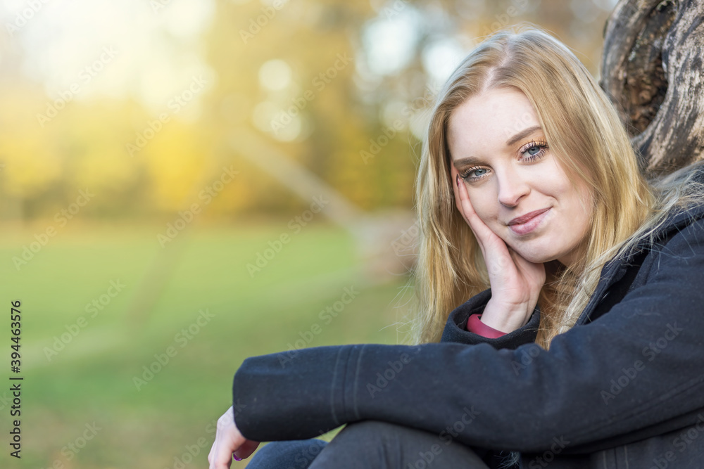 Smiling young woman is posing in the autumn park supporting her head with hand looking at the camera. Horizontally. Smiling young woman is posing in the autumn park supporting her head with hand looking at the camera. Horizontally.
