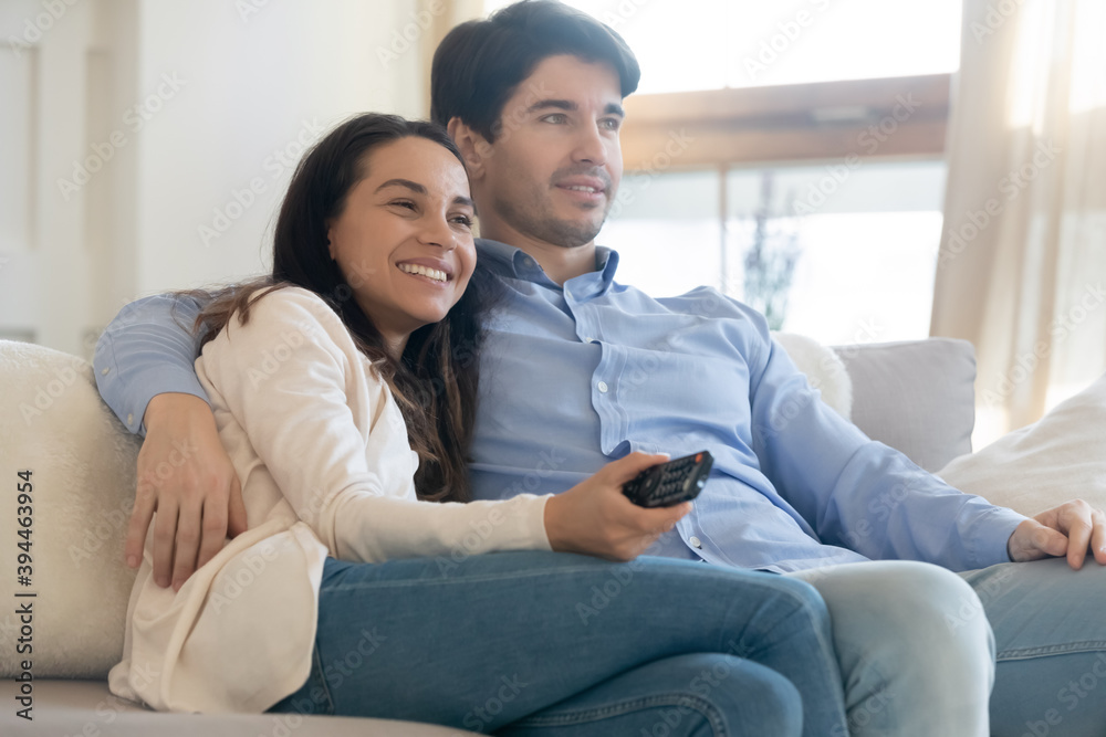 Relaxing by screen. Happy young spouses or couple in love cuddling on soft couch at home resting from work watching favorite movie program by satellite cable tv choosing channel using remote control Relaxing by screen. Happy young spouses or couple in love cuddling on soft couch at home resting from work watching favorite movie program by satellite cable tv choosing channel using remote control