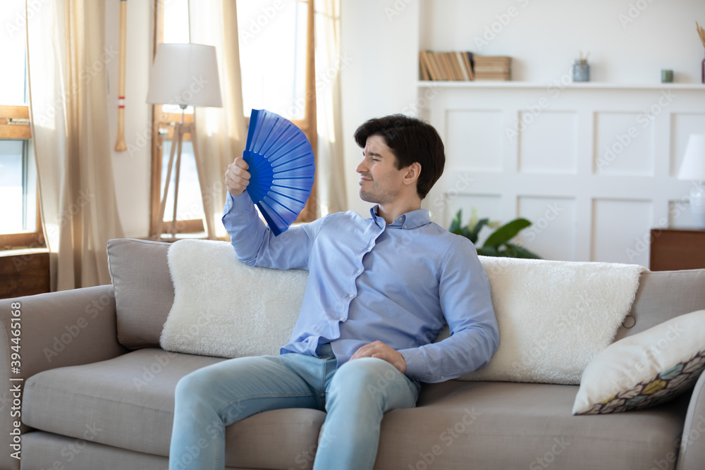 Overheated young man owner renter tenant of house flat resting on sofa in living room saving himself from heat using paper weaver understanding problem of bad ventilation and lack of air conditioning. Overheated young man owner renter tenant of house flat resting on sofa in living room saving himself from heat using paper weaver understanding problem of bad ventilation and lack of air conditioning.