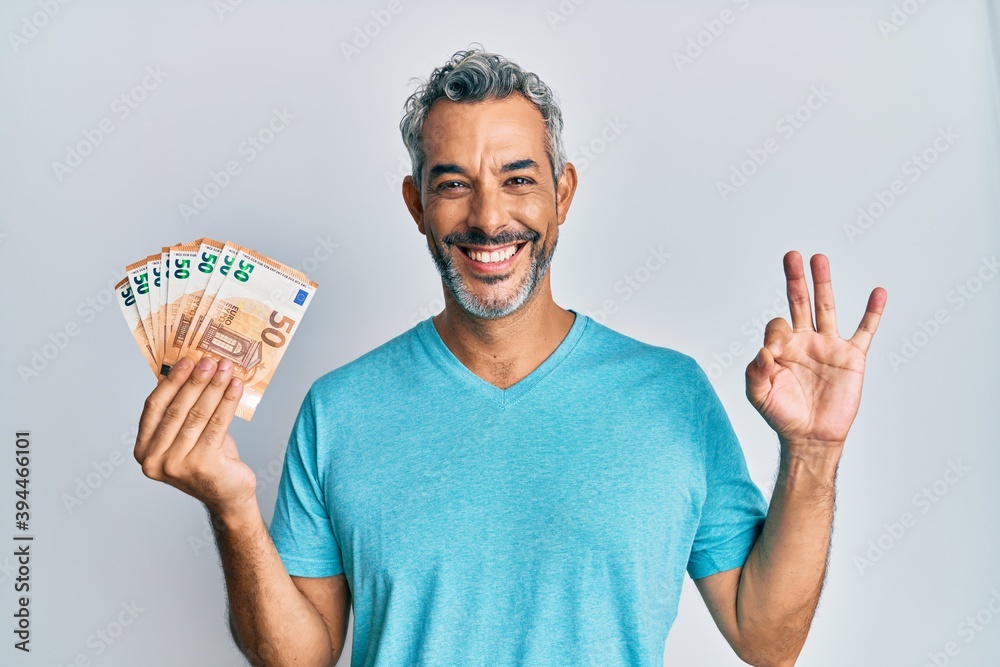 Middle age grey-haired man holding 50 euro banknotes doing ok sign with fingers, smiling friendly gesturing excellent symbol Middle age grey-haired man holding 50 euro banknotes doing ok sign with fingers, smiling friendly gesturing excellent symbol