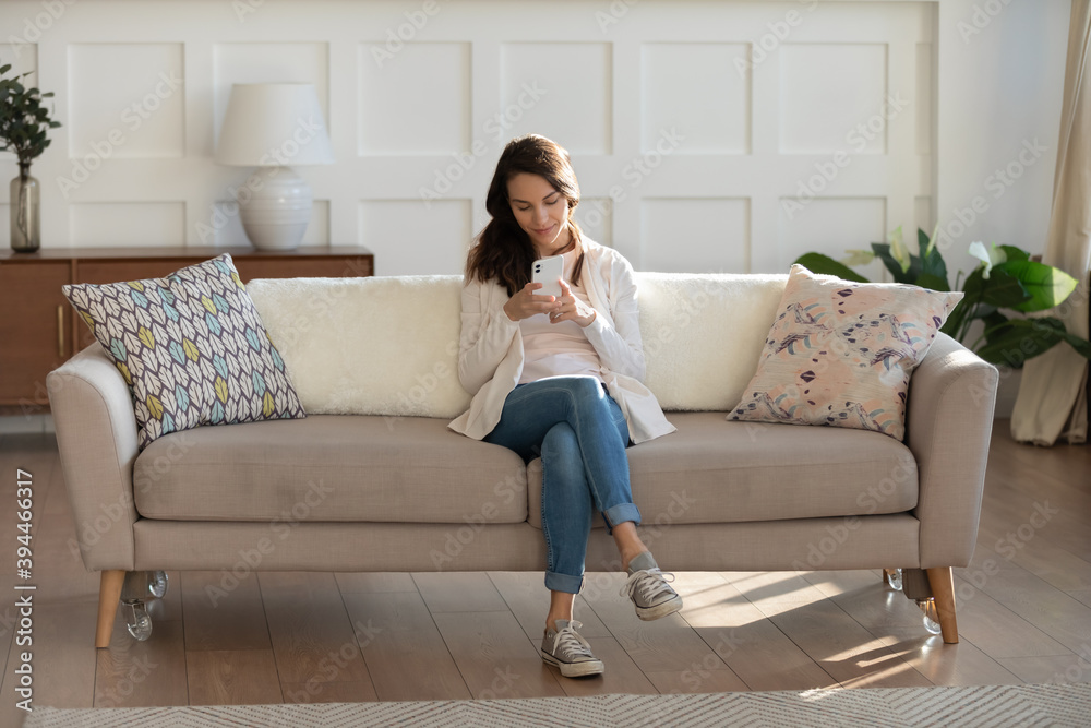 Online activity. Calm smiling young woman owner renter tenant of house apartment flat sitting on couch in modern living room using mobile phone chatting shopping making video call reading good news Online activity. Calm smiling young woman owner renter tenant of house apartment flat sitting on couch in modern living room using mobile phone chatting shopping making video call reading good news