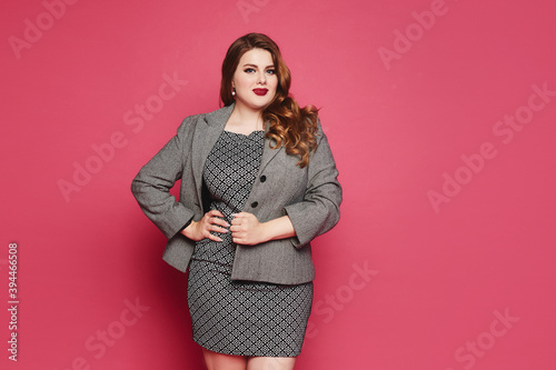 Charming plus size businesswoman wearing a grey jacket and dress looking at the camera and standing at pink background, isolated. XXL model in official outfit posing over pink