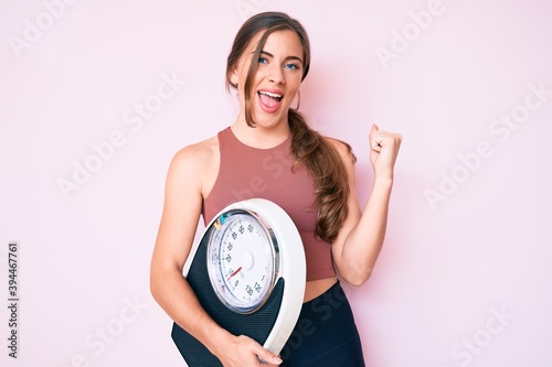 Beautiful young caucasian woman holding weight machine to balance weight loss screaming proud, celebrating victory and success very excited with raised arms