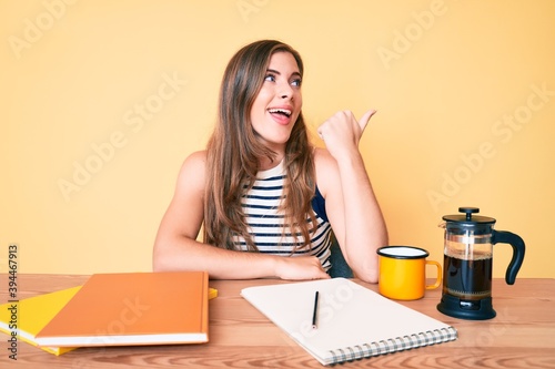 Beautiful young caucasian woman sitting on the table stuying for university pointing thumb up to the side smiling happy with open mouth photo