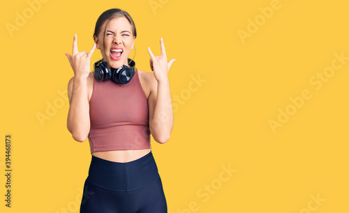 Beautiful caucasian young woman wearing gym clothes and using headphones shouting with crazy expression doing rock symbol with hands up. music star. heavy music concept.