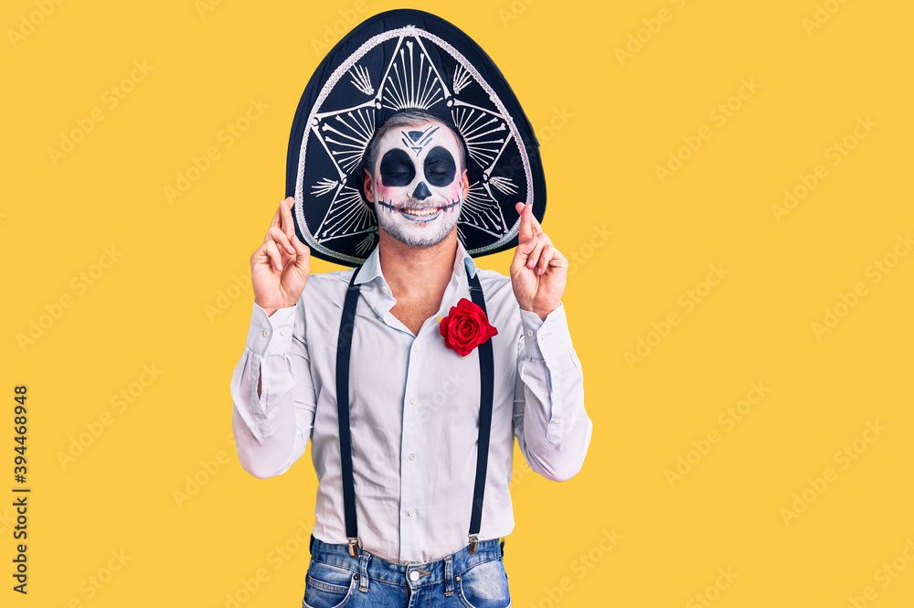 Man wearing day of the dead costume over background gesturing finger crossed smiling with hope and eyes closed. luck and superstitious concept. Man wearing day of the dead costume over background gesturing finger crossed smiling with hope and eyes closed. luck and superstitious concept.