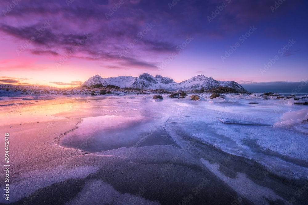 Frozen sea coast at colorful sunset in Lofoten islands, Norway. Snowy mountains, sea with frosty shore, ice, reflection in water, purple sky . Winter landscape with snow covered rocks, fjord at night