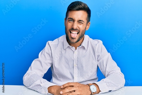 Handsome hispanic man wearing business clothes sitting on the table sticking tongue out happy with funny expression. emotion concept. photo