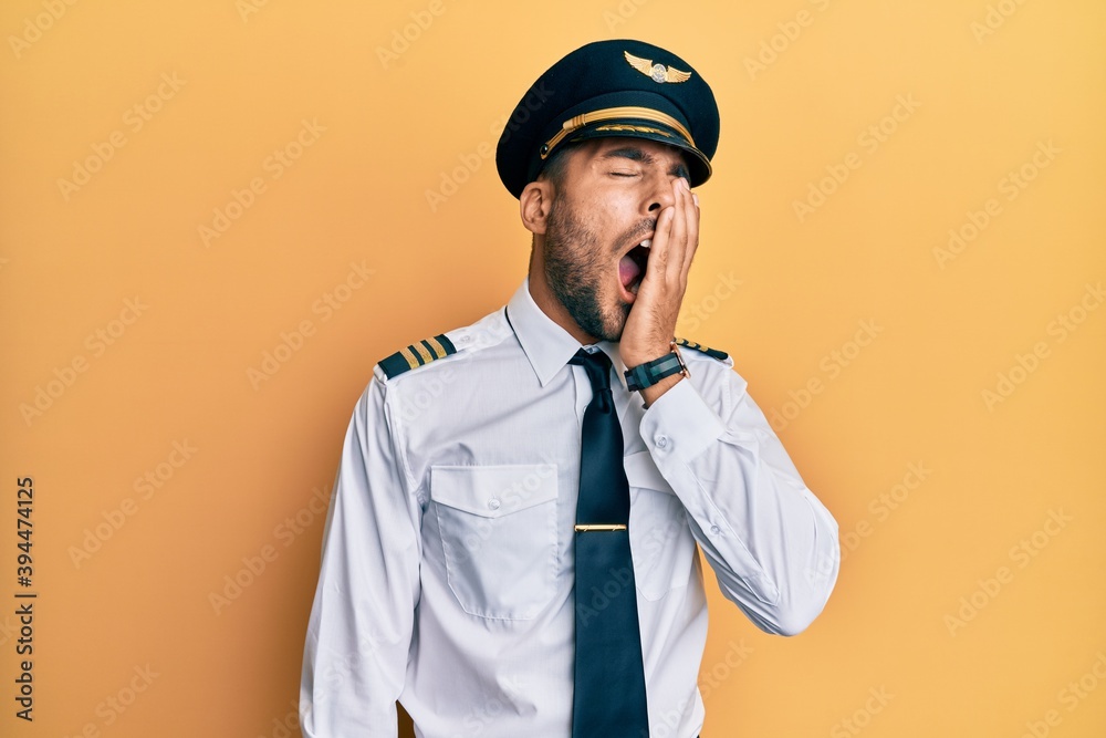 Handsome hispanic man wearing airplane pilot uniform yawning tired covering half face, eye and mouth with hand. face hurts in pain.