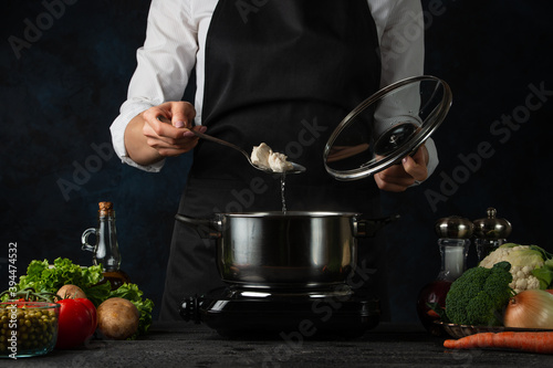 Chef in black apron testing hot soup at the professional restaurant kitchen on dark blue background. Backstage of preparing dish for dinner. Coking process concept. Cookbook.