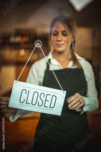 Portrait of a Caucasian woman with a black apron, standing behind a glass door with the sign ''closed''