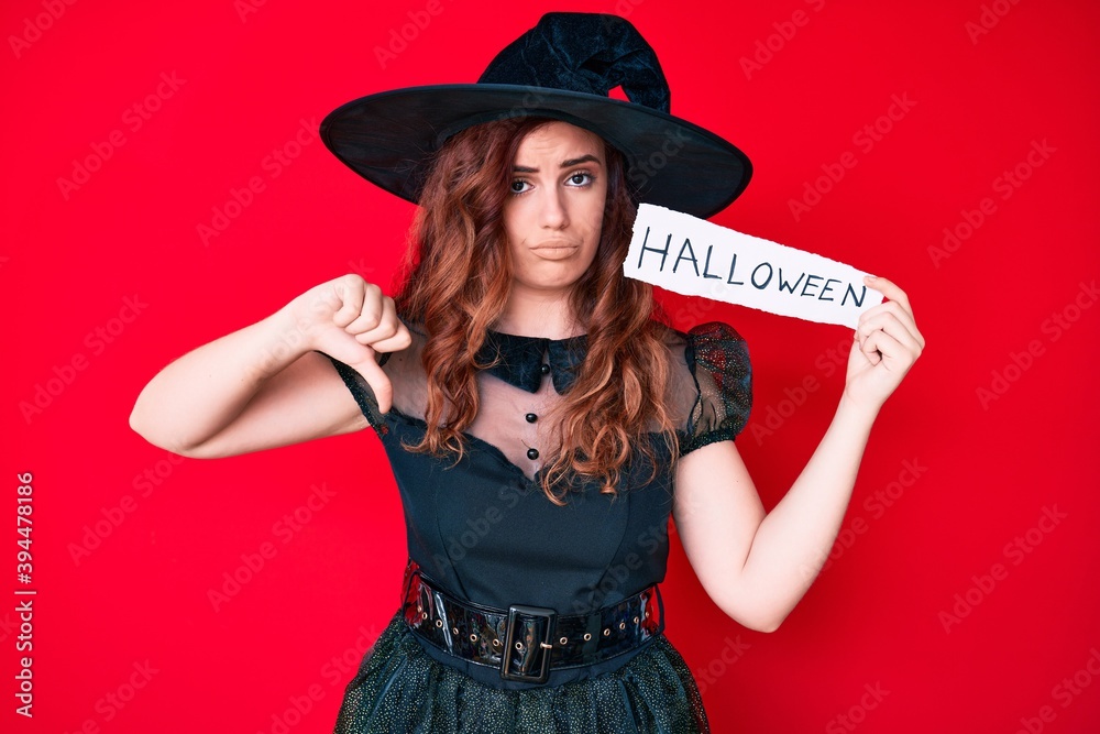 Young beautiful woman wearing witch costume holding paper with halloween message with angry face, negative sign showing dislike with thumbs down, rejection concept