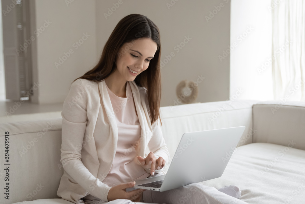 Working from home. Calm smiling young woman sitting on sofa with laptop on knees concentrated on work study online, shopping in internet store, chatting with friends using messenger app, sending email Working from home. Calm smiling young woman sitting on sofa with laptop on knees concentrated on work study online, shopping in internet store, chatting with friends using messenger app, sending email
