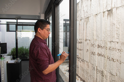 A latin man with glasses, probably an architect or a entrepreneur writing in a glass door in a office, dressing informal clothes 