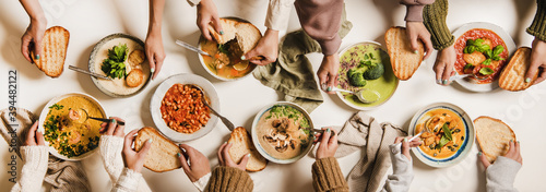 People eating Autumn and Winter creamy vegan soups. Flat-lay of peoples hands, soup plates and bread slices over plain white table background, top view. Fall and Winter food menu, vegetarian food