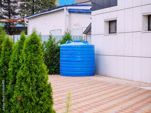 A blue plastic water tank stands in a backyard surrounded by thujas photo