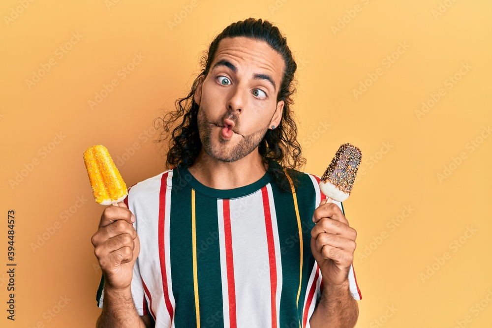 Young handsome man with long hair eating two ice cream making fish face with mouth and squinting eyes, crazy and comical. Young handsome man with long hair eating two ice cream making fish face with mouth and squinting eyes, crazy and comical.