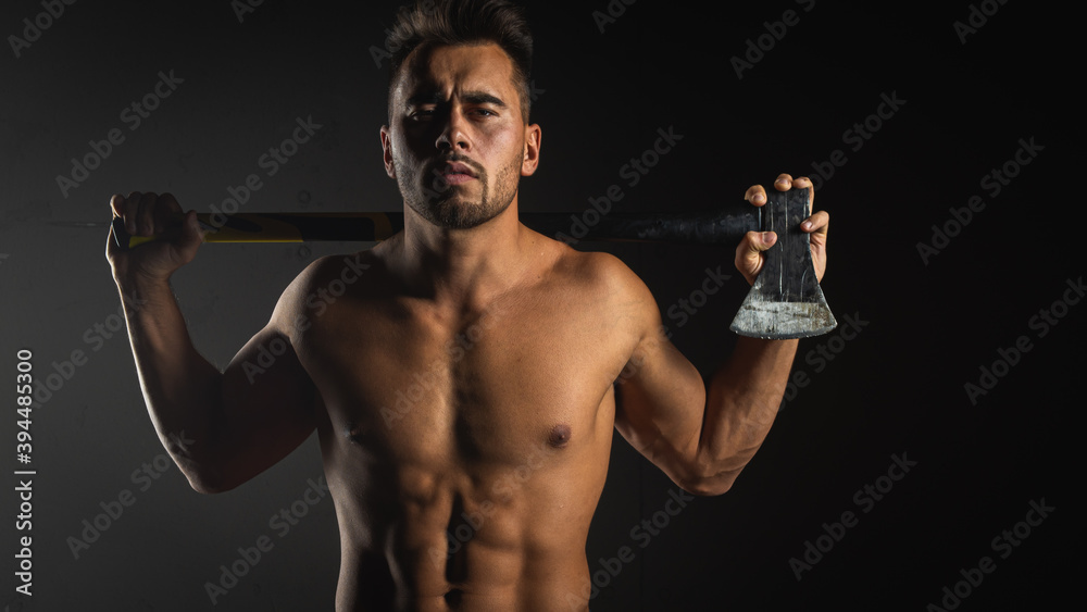 young handsome adult, muscular firefighter in uniform holding ax fire equipment in hands, pensive, isolated on dark background. Low key