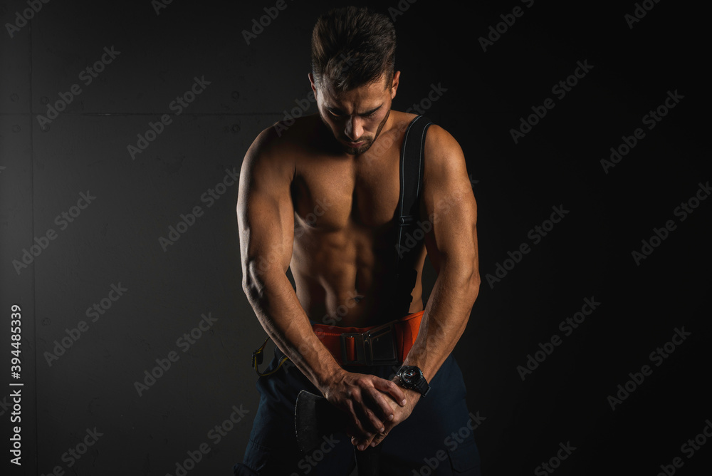 young, handsome, adult, muscular firefighter in uniform holding his head bowed and leaning on the fire ax that is in front of him, isolated on a dark background. Low key. black and white photo
