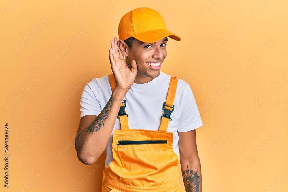 Young handsome african american man wearing handyman uniform over yellow background smiling with hand over ear listening an hearing to rumor or gossip. deafness concept. Young handsome african american man wearing handyman uniform over yellow background smiling with hand over ear listening an hearing to rumor or gossip. deafness concept.