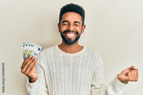 Handsome young hispanic man holding bunch of 5 euro banknotes celebrating achievement with happy smile and winner expression with raised hand