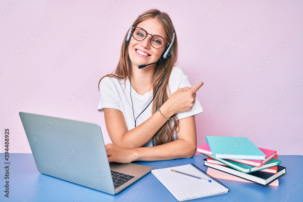 Young blonde girl wearing operator headset at the call center office smiling cheerful pointing with hand and finger up to the side