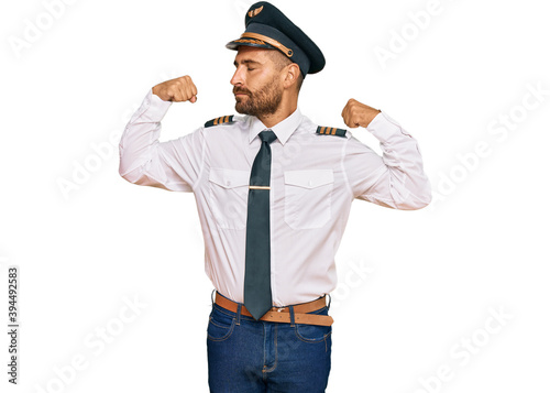 Handsome man with beard wearing airplane pilot uniform showing arms muscles smiling proud. fitness concept.