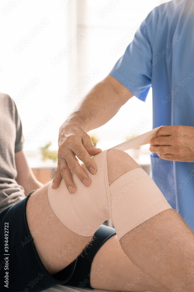 Obraz premium Hands of clinician in blue uniform wrapping injured knee of young patient