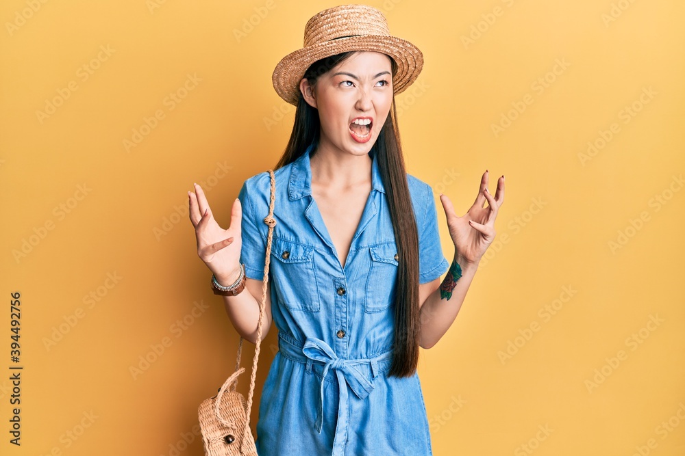 Young chinese woman wearing summer hat crazy and mad shouting and yelling with aggressive expression and arms raised. frustration concept. Young chinese woman wearing summer hat crazy and mad shouting and yelling with aggressive expression and arms raised. frustration concept.