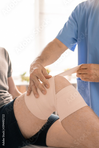Hands of clinician in blue uniform wrapping injured knee of young patient