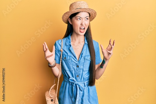 Young chinese woman wearing summer hat crazy and mad shouting and yelling with aggressive expression and arms raised. frustration concept.