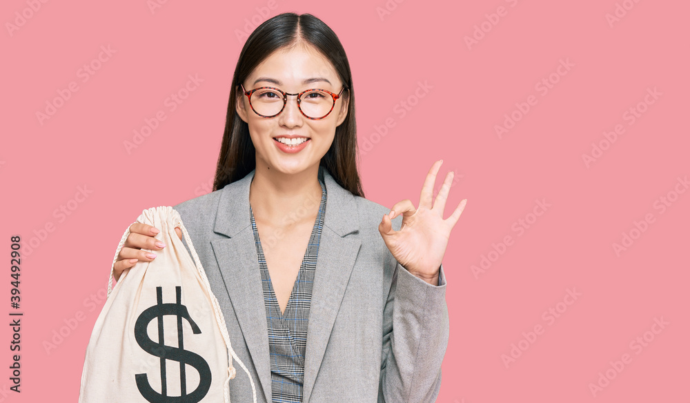 Young chinese woman wearing business suit holding dollars bag doing ok sign with fingers, smiling friendly gesturing excellent symbol Young chinese woman wearing business suit holding dollars bag doing ok sign with fingers, smiling friendly gesturing excellent symbol