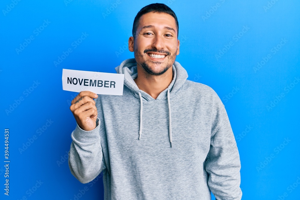 Handsome man with tattoos holding november word on paper looking positive and happy standing and smiling with a confident smile showing teeth Handsome man with tattoos holding november word on paper looking positive and happy standing and smiling with a confident smile showing teeth