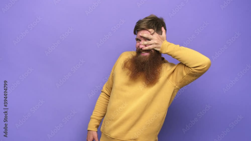 Young adult bearded caucasian man isolated blink at the camera through fingers, embarrassed covering face Young adult bearded caucasian man isolated blink at the camera through fingers, embarrassed covering face
