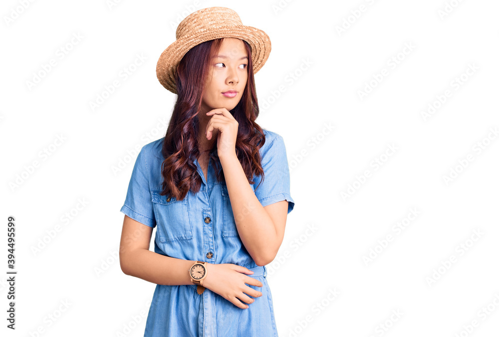 Young beautiful chinese girl wearing summer hat with hand on chin thinking about question, pensive expression. smiling with thoughtful face. doubt concept. Young beautiful chinese girl wearing summer hat with hand on chin thinking about question, pensive expression. smiling with thoughtful face. doubt concept.