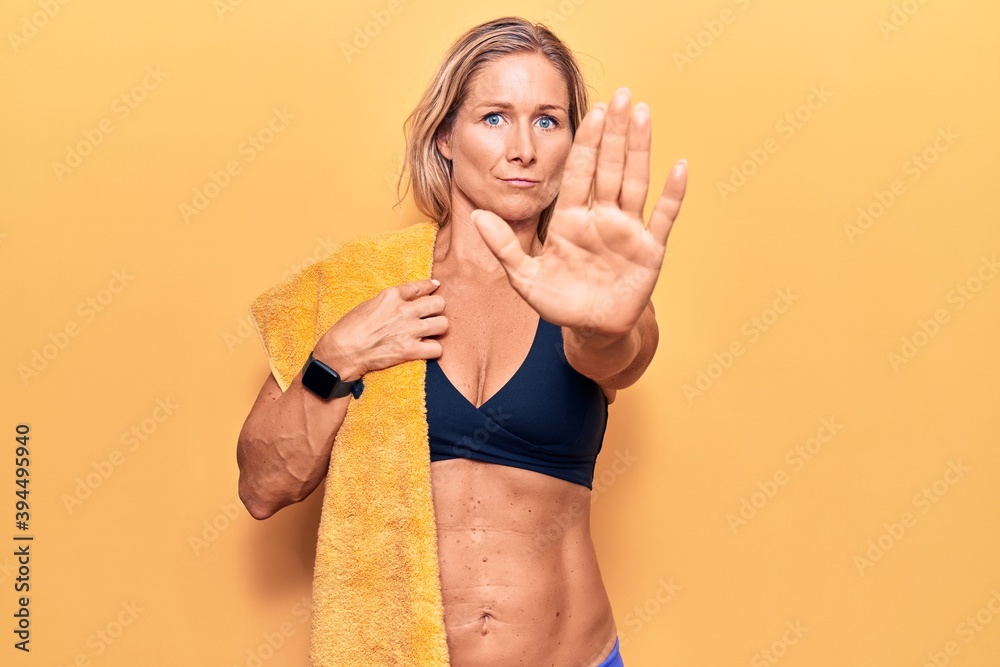 Middle age caucasian blonde woman wearing bikini and towel with open hand doing stop sign with serious and confident expression, defense gesture Middle age caucasian blonde woman wearing bikini and towel with open hand doing stop sign with serious and confident expression, defense gesture