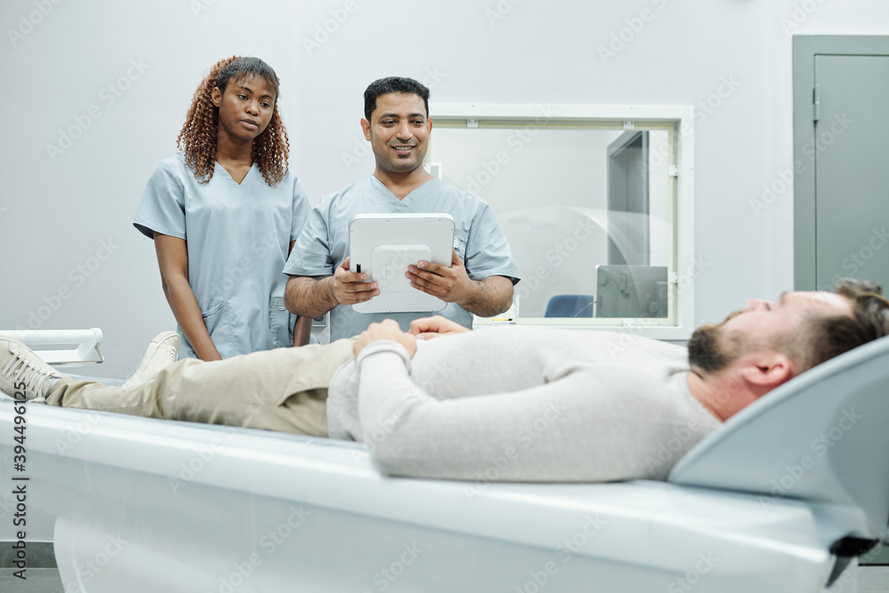 Young assistant and doctor in uniform standing by long couchette with patient Young assistant and doctor in uniform standing by long couchette with patient